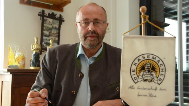 Ein Mann mit Brille und Trachtenjacke sitzt vor einem Tisch mit einem Wimpel der Käserei „Die Käsemacher“.