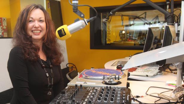 Eine Frau lächelt in einem Radiostudio neben einem Mischpult und einem Mikrofon.
