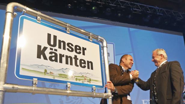 Zwei Männer schütteln sich vor einem Schild mit der Aufschrift „Unser Kärnten“ die Hände.