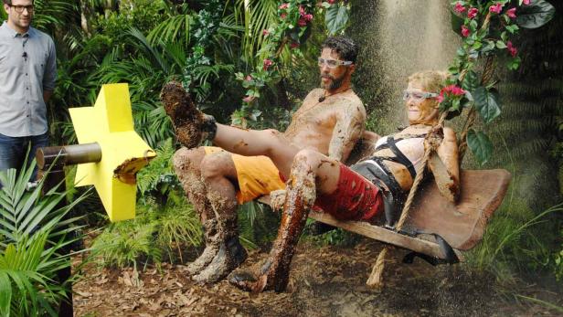 Zwei schlammbedeckte Personen sitzen auf einer Schaukel im Dschungel.