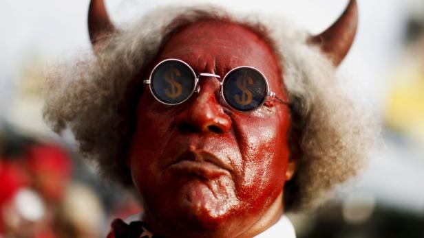 Eine Person mit roter Farbe im Gesicht, Hörnern und Brille mit Dollarzeichen.