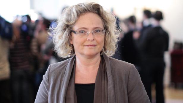 Eine Frau mit blond gelocktem Haar und Brille lächelt in die Kamera.