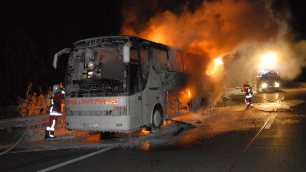 Ein brennender Reisebus auf einer Autobahn bei Nacht, mit Feuerwehrleuten im Einsatz.
