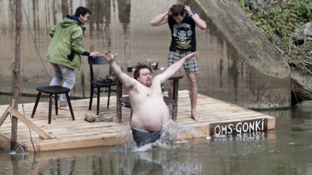 Ein Mann fällt von einem Holzfloß ins Wasser, während zwei andere zusehen.