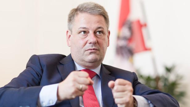 Ein Mann im Anzug sitzt mit geballten Fäusten vor einer österreichischen Flagge.