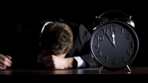 Desperate businessman in dark suit sitting at office desk with head down being in despair with close up of alarm clock in foreground showing five minutes to twelve o-c&#039;clock, low-key style isolated on black background.
