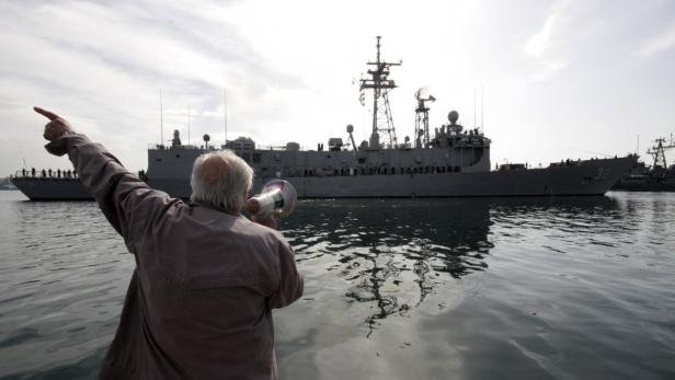 Ein Mann mit Megafon zeigt auf ein Kriegsschiff im Hafen.