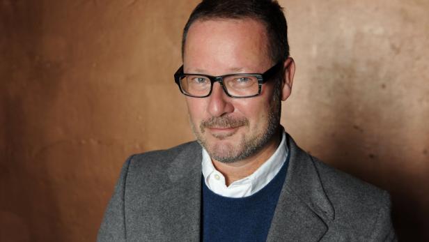 Ein Mann mit Brille, Bart und grauem Sakko vor einem braunen Hintergrund.