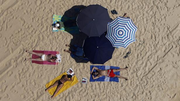 Luftaufnahme von Menschen, die sich am Strand unter Sonnenschirmen sonnen.