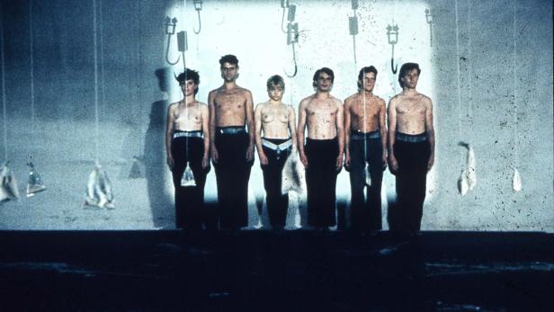 Sechs barbusige Personen stehen vor einer Wand mit hängenden Objekten.