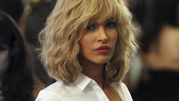 Eine Frau mit blonden, gewellten Haaren und einem weißen Hemd blickt in die Kamera.