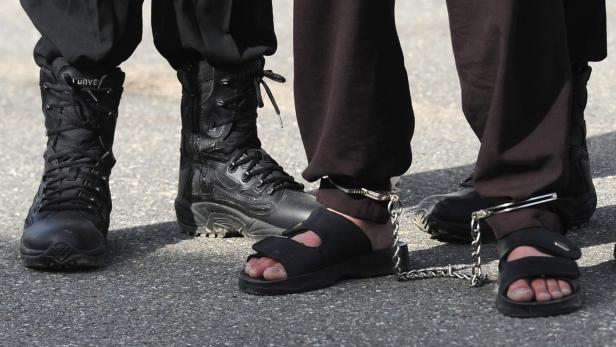 Eine Person in Handschellen und Sandalen steht neben Personen in Stiefeln.