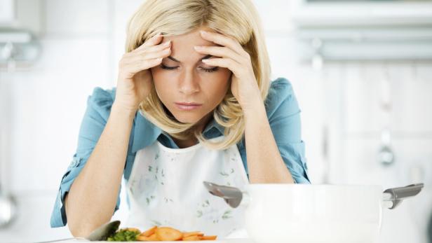 Eine Frau mit Kopfschmerzen sitzt in der Küche vor einem Topf und Gemüse.