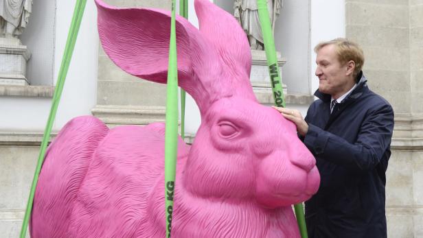 Kopie von WIEN: GROSSE DÜRER-HASEN-SKULPTUR PER KRAN AUF DEN Ein Mann berührt eine riesige, pinkfarbene Hasenskulptur, die mit grünen Gurten befestigt ist.