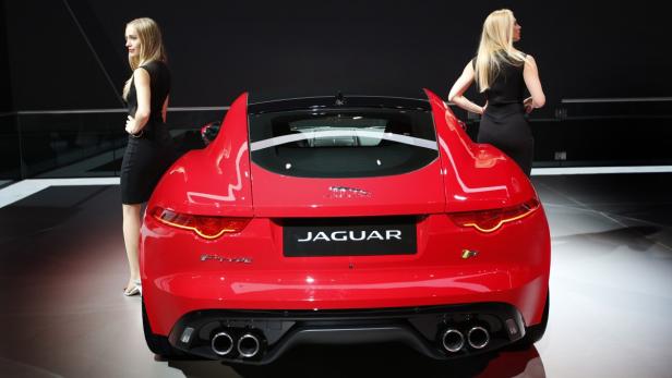 Ein roter Jaguar F-Type wird von zwei Frauen in schwarzen Kleidern flankiert.