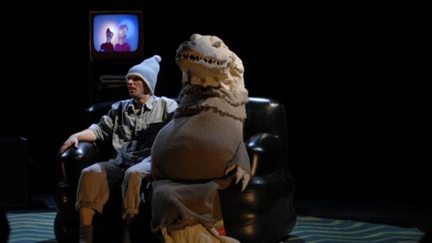 Ein Mann mit Mütze sitzt neben einem Dinosaurier-Kostüm auf einem Sessel und schaut fern.