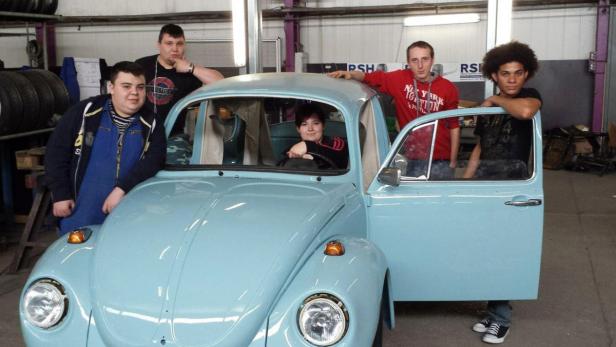 Eine Gruppe von Personen posiert mit einem hellblauen VW Käfer in einer Werkstatt.