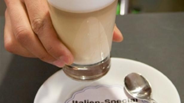 Ein Latte Macchiato mit der Aufschrift „Pisa 246€“ wird auf einer Untertasse mit Expedia-Werbung serviert.