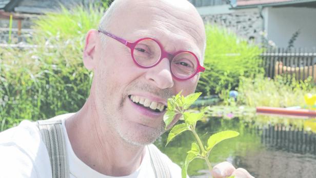 Ein Mann mit Brille hält eine Pflanze vor einem Teich in die Kamera.