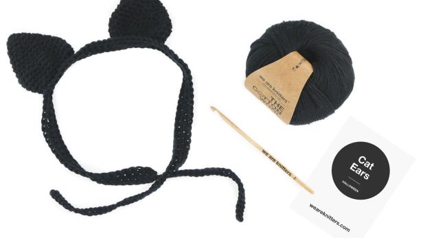 Ein Stirnband mit gehäkelten Katzenohren, eine Häkelnadel und ein Wollknäuel von We Are Knitters.