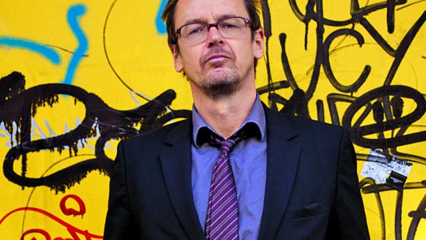 Ein Mann mit Brille und Anzug steht vor einer gelben Wand mit Graffiti.