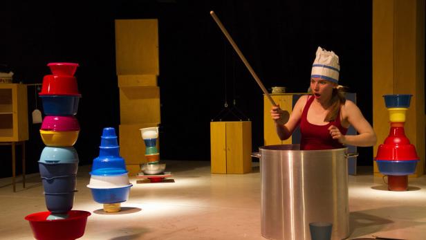 Eine Frau mit Kochmütze steht in einem großen Topf und hält einen Kochlöffel.