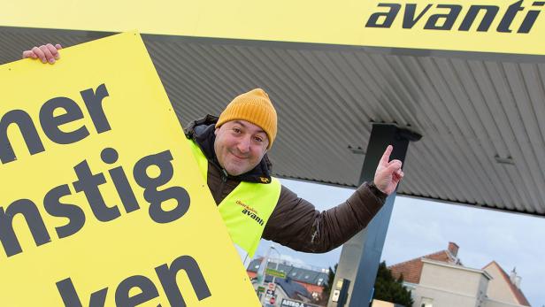 Ein Mann mit gelber Mütze hält ein Schild mit der Aufschrift „Immer günstiger tanken“ vor einer Avanti-Tankstelle.