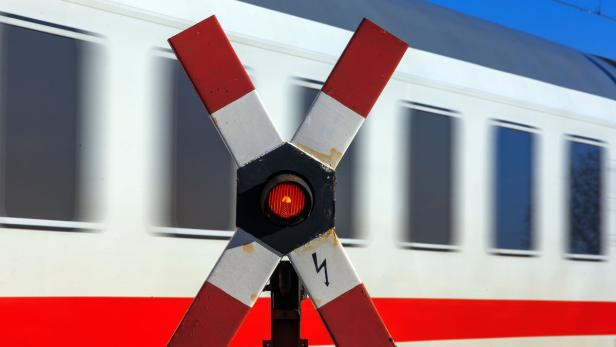 Ein Andreaskreuz mit rotem Blinklicht vor einem vorbeifahrenden Zug.