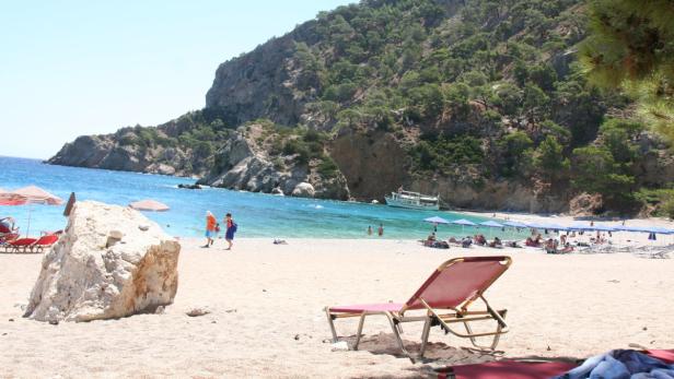 Ein Strand mit Liegestühlen, Badegästen und einem bewaldeten Hügel im Hintergrund.