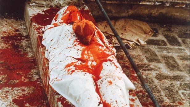 Eine in ein weißes Tuch gehüllte Figur liegt auf einem Steinaltar, bedeckt mit roter Farbe.