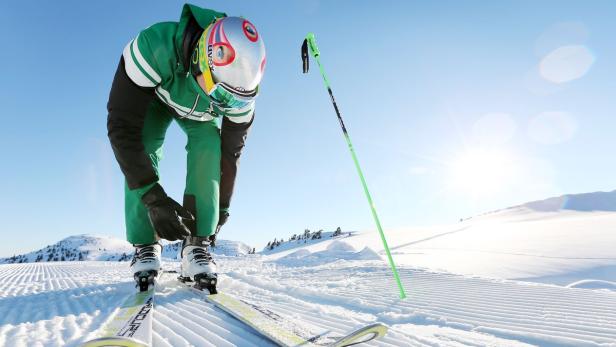 Ein Skifahrer in grüner Kleidung schnallt seine Skischuhe auf einer Skipiste fest.