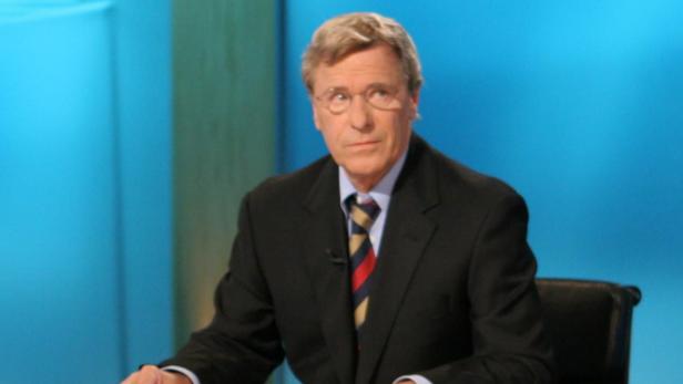 Ein Mann mit Brille und Anzug sitzt vor einem blauen Hintergrund.