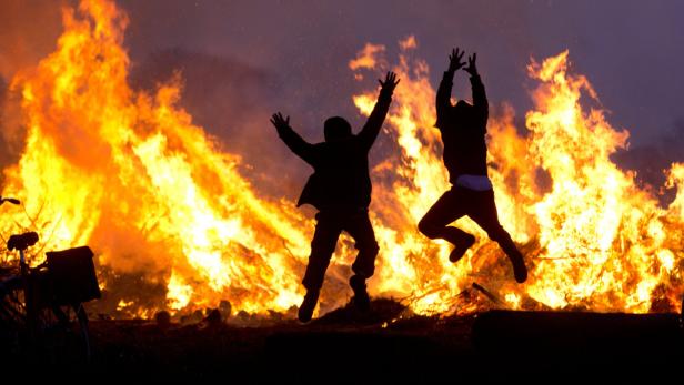 Zwei Personen springen vor einem großen Osterfeuer in die Luft.