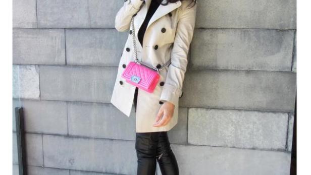 Eine Frau in einem Trenchcoat, Leggings und pinkfarbenen High Heels posiert mit einer pinkfarbenen Handtasche vor einer grauen Wand.
