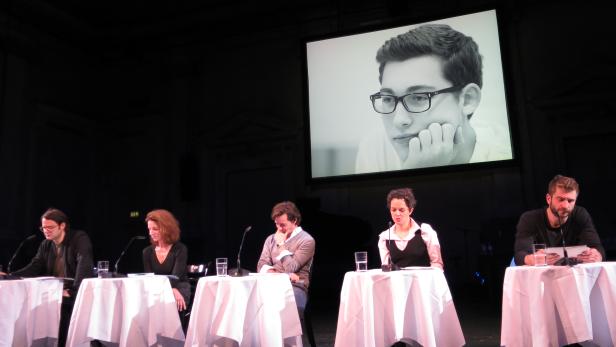 Fünf Personen sitzen an Tischen auf einer Bühne vor einer Leinwand mit einem Porträt.