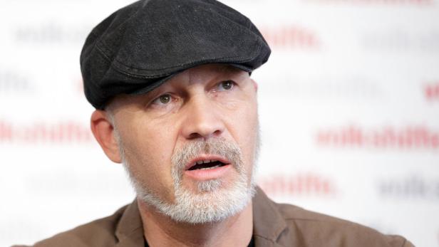 Ein Mann mit Bart und Schiebermütze spricht vor einer Wand mit Logos.