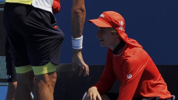 Ein Balljunge in roter Kleidung kniet neben einem Tennisspieler auf dem Platz.