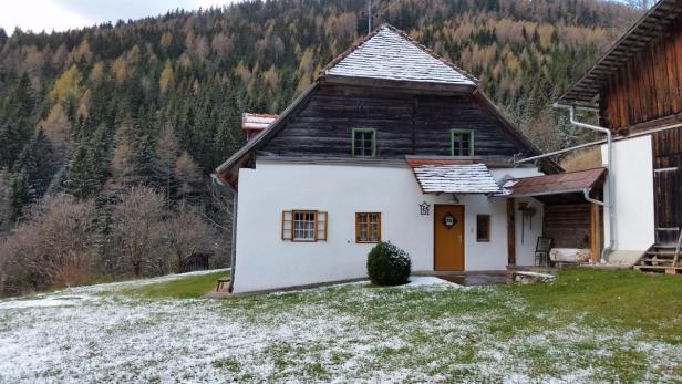 Ein traditionelles Bauernhaus mit Schnee bedecktem Dach vor einem bewaldeten Hügel.
