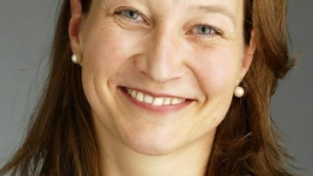 Eine lächelnde Frau mit braunen Haaren und Perlenohrringen vor einem grauen Hintergrund.