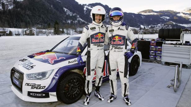 Zwei Rennfahrer posieren vor einem Audi-Rennwagen mit Red Bull- und Need for Speed-Logos.