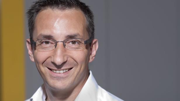 Ein lächelnder Mann mit Brille und weißem Hemd vor einem grauen Hintergrund.