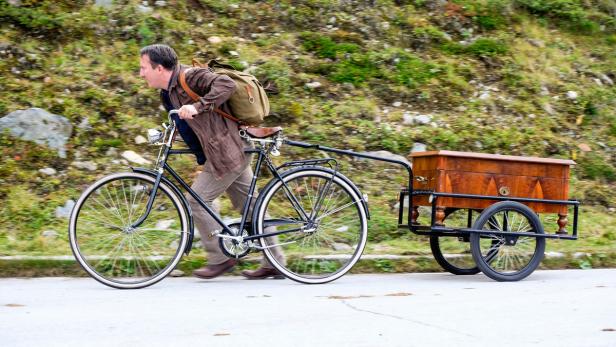 Ein Mann schiebt sein Fahrrad, an dem ein Anhänger mit einer Holzkiste befestigt ist.
