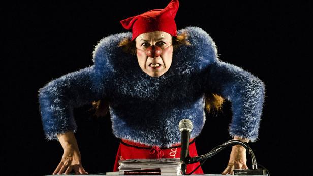 Eine Frau mit roter Nase und Zipfelmütze steht wütend an einem Rednerpult.