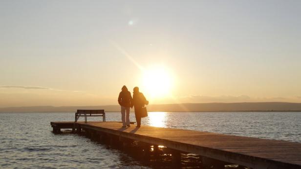 Zwei Personen gehen bei Sonnenuntergang auf einem Steg am Wasser entlang.