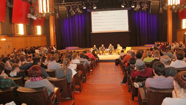 Publikum sitzt in einem Hörsaal und verfolgt eine Podiumsdiskussion mit Yilmaz Gülüm und anderen.