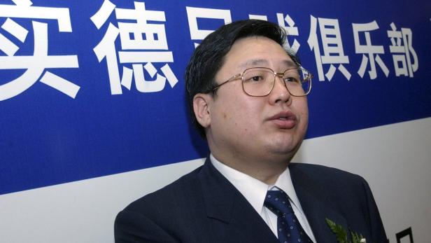 Ein Mann mit Brille und Anzug steht vor einem blauen Hintergrund mit chinesischen Schriftzeichen.