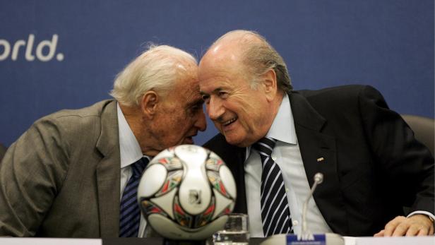 Zwei Männer in Anzügen stehen an einem Tisch mit einem Fußball.