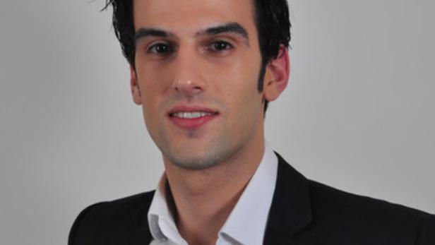 Ein Mann mit dunklen Haaren und einem schwarzen Anzug lächelt in die Kamera.