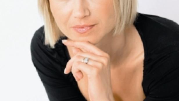 Eine Frau mit blonden Haaren und schwarzem Oberteil posiert mit der Hand am Kinn.