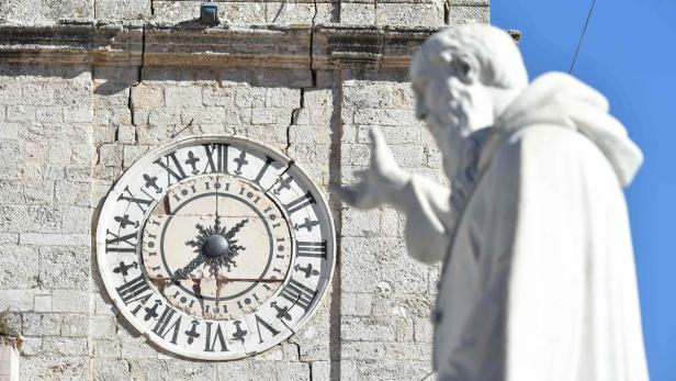 Eine Uhr mit römischen Ziffern an einer steinernen Gebäudefassade, neben einer Statue.